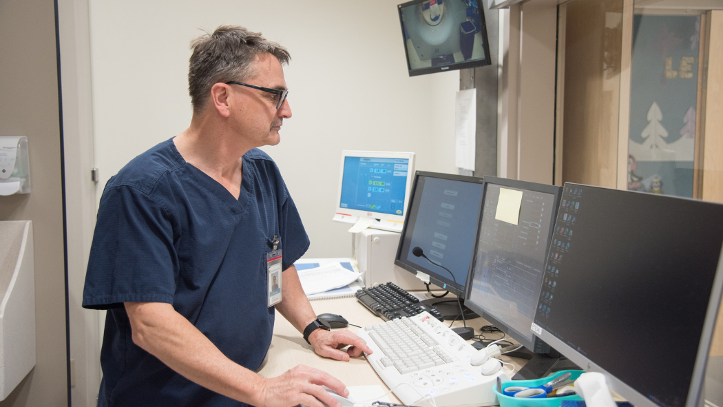 A person in scrubs and glasses works at a desk with multiple computer monitors and medical equipment in an office setting.