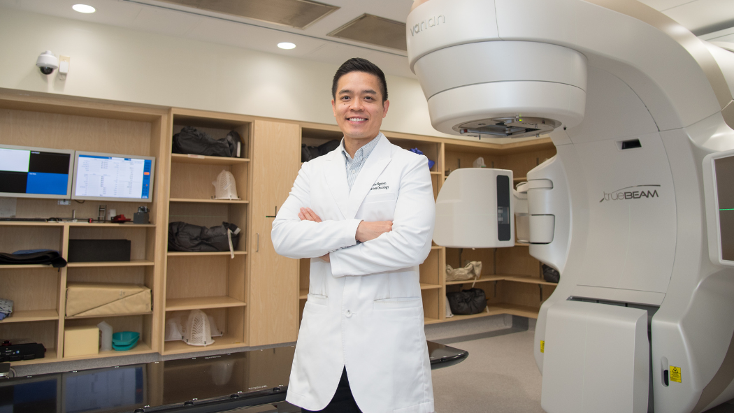 A person in a white coat stands confidently beside a large medical machine in a modern clinical setting.