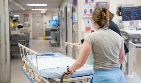 A healthcare worker pushes an empty hospital bed down a corridor, past medical equipment and open doors, in a brightly lit hospital.