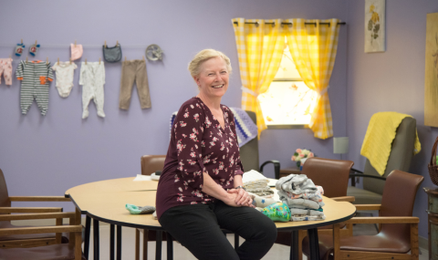 Deb Lines, Recreational Therapist smiling while leaning against table in a lilac purple coloured room