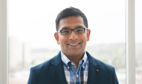 A person wearing glasses and a blue blazer stands in front of a window with a blurred outdoor background.