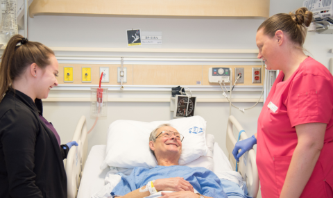 An elderly patient in a hospital bed smiles at two medical staff members, one in black and the other in red scrubs, who stand beside him.