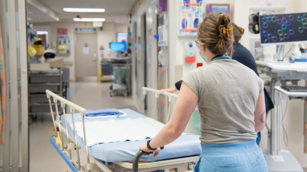A healthcare worker pushes an empty hospital bed down a corridor, past medical equipment and open doors, in a brightly lit hospital.