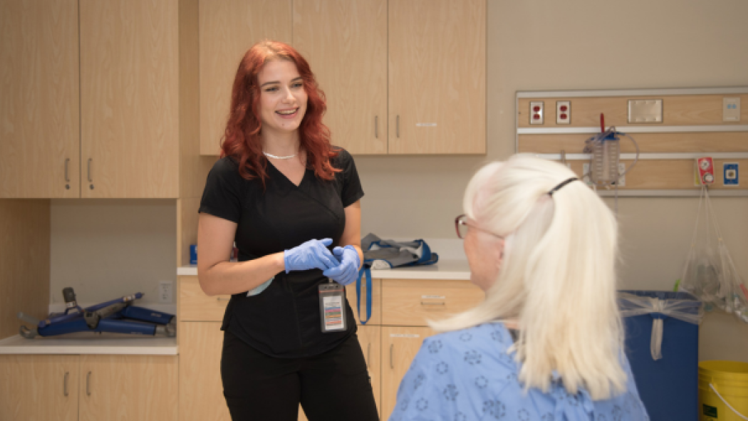 A health-care professional with red hair speaks to a patient in a medical examination room.