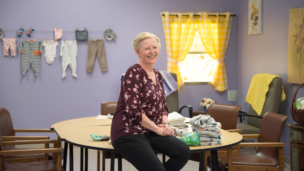 Deb Lines, Recreational Therapist smiling while leaning against table in a lilac purple coloured room