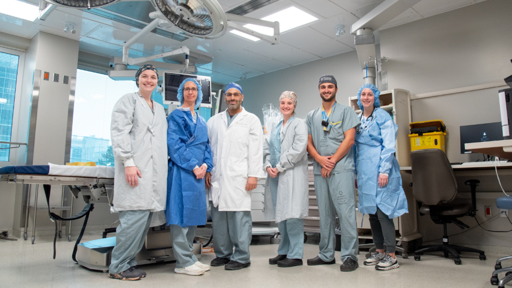 A group of six medical professionals in scrubs and lab coats stand together in a hospital room with medical equipment.