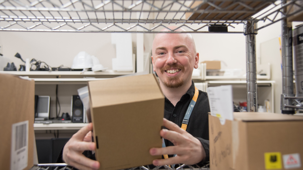 Team member smiles at camera while reaching for box of IT equipment from shelf